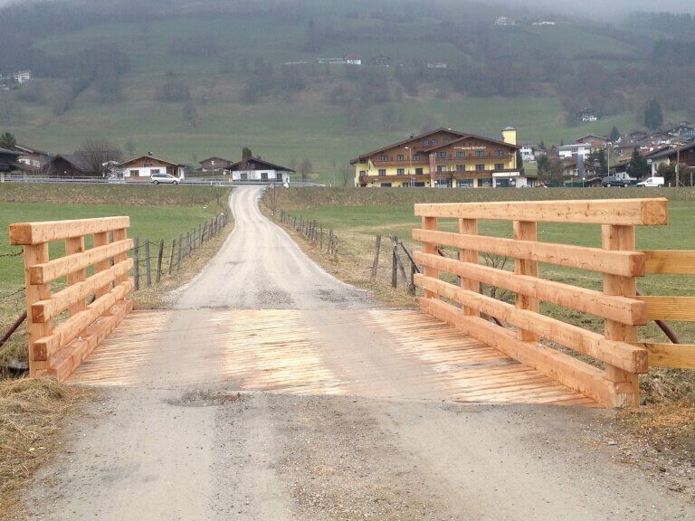 Holzbrücke über einen Feldweg, umgeben von Wiesen und Gebäuden im Pinzgau.