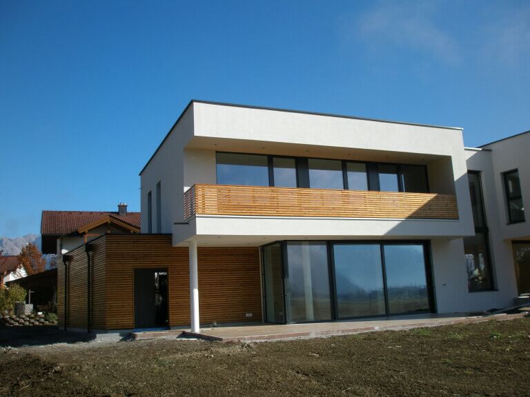 Modernes Holzhaus mit großem Fensterfront und Balkon, umgeben von Gartenfläche.